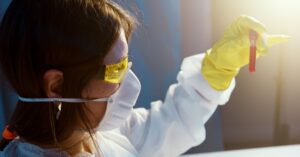 Cientista com óculos de proteção e luvas segurando um tubo de ensaio vermelho em um ambiente de laboratório.