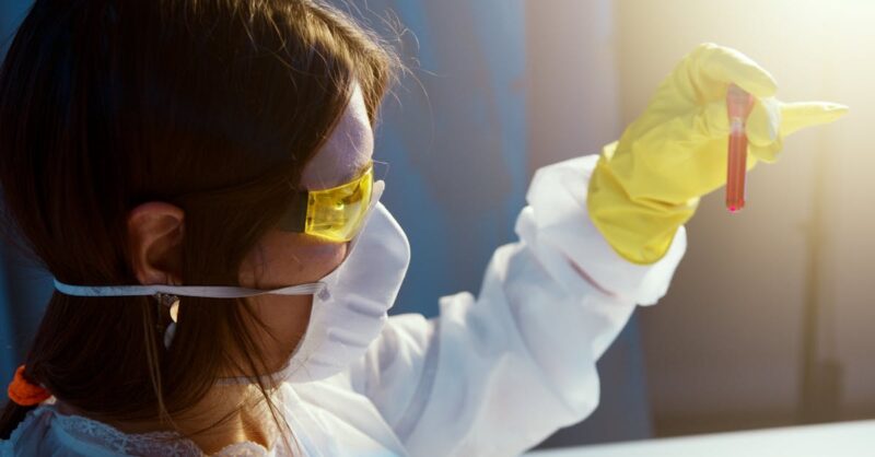 Cientista com óculos de proteção e luvas segurando um tubo de ensaio vermelho em um ambiente de laboratório.