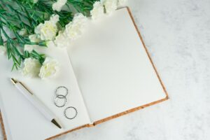 Caderno aberto em página em branco com caneta, alianças de prata e cravos brancos sobre superfície de mármore