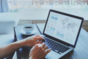 Mãos digitando em notebook que exibe painel de métricas com gráficos e mapa-múndi, ao lado de uma xícara de café