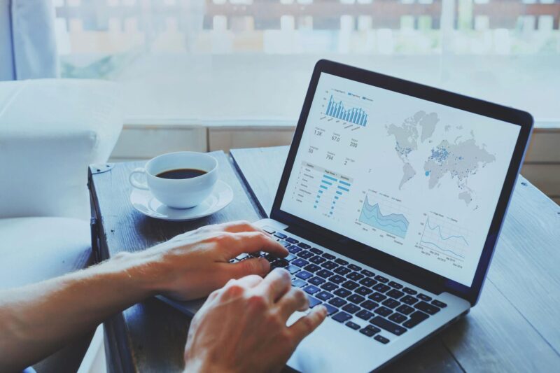 Mãos digitando em notebook que exibe painel de métricas com gráficos e mapa-múndi, ao lado de uma xícara de café